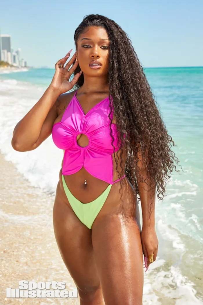 Megan Thee Stallion stands on the sand in a green and pink floral one-piece swimsuit.